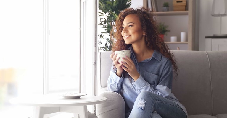 Smiling woman sat on stylish sofa drinking coffee looking out of window