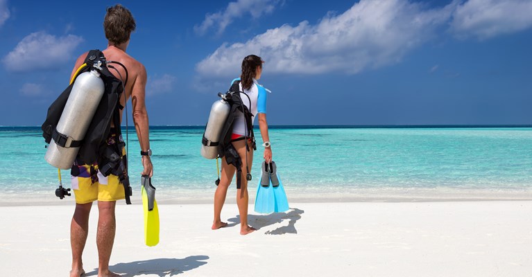 Two young people walking on the white sandy beach towards the crystal blue sea with scuba diving kit in hand