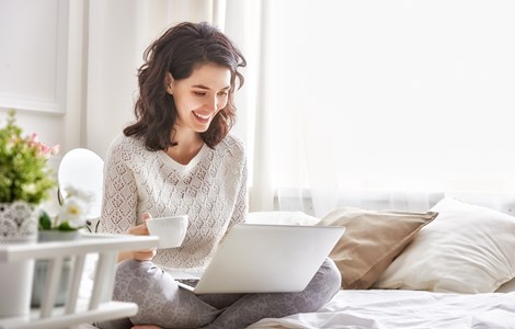 Young woman smiling with drink and laptop sat on bed