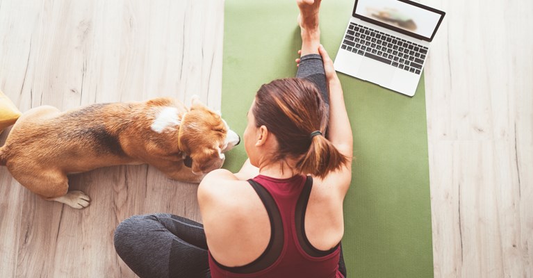 Woman doing yoga on floor with laptop and dog beside her