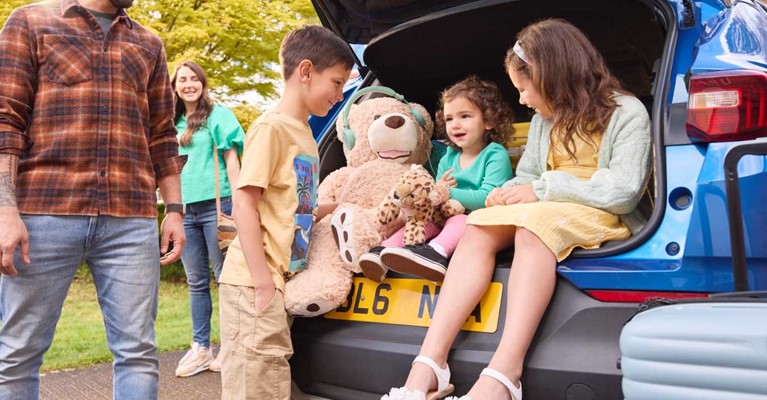 Young children sit in the open boot of a car while parents look on