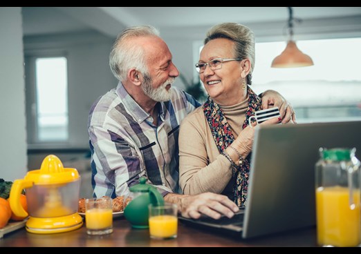 Older couple smile at each other using laptop to browse financial services in their home