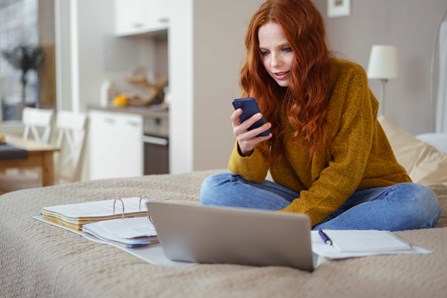 Woman in yellow jumper reviewing financial services on mobile and laptop in her home