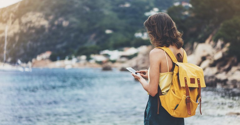 Girl in summer clothes with yellow backpack checking phone by the sea