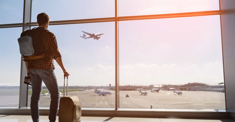 Man with backpack and wheel suitcase watches plane take off from airport window