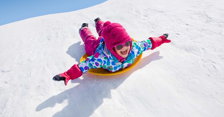 little girl in pink hat and goggles sliding down a snow slope on a sled
