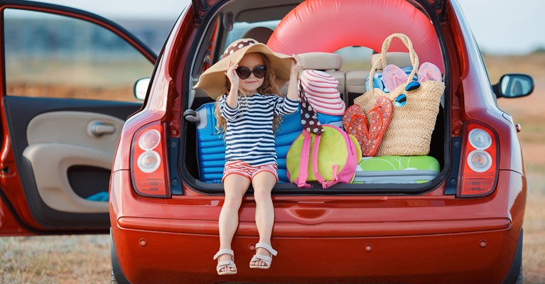 Little girl in a blue dress sat in the boot of a small red car with beach accessories on a sandy beach