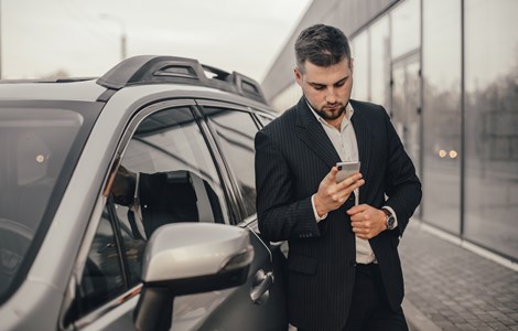 Young man in a suit standing by a silver car scrolling through his phone in a showroom looking for car insurance