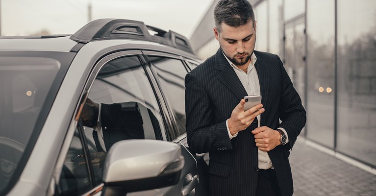 Young man in a suit standing by a silver car scrolling through his phone in a showroom looking for car insurance