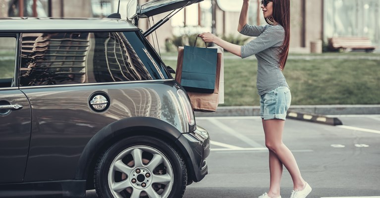 Young girl wearing summer clothes loading paper shopping bags into the boot of her small grey car