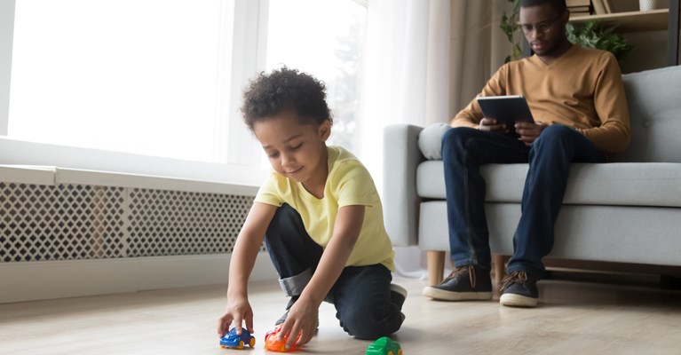 Young boy playing with his toys on the floor in the front room whilst his dad is sat on the sofa behind him on his tablet