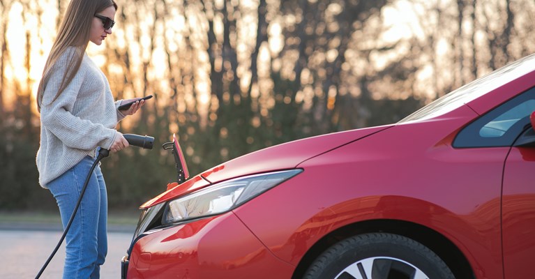 Young girl with sunglasses on charging her red electric car whilst looking at her mobile phone 