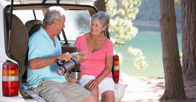 Couple sat in the boot of their silver car enjoying some snacks and coffee from a flask whilst parked up by a lake