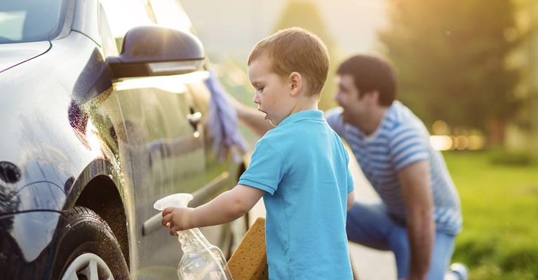 Young dad cleaning his small blue car with young son outside their house 