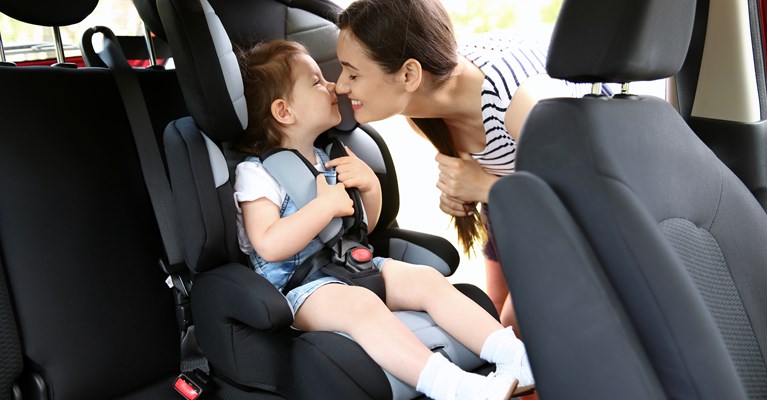 Young daughter sat in a car seat in the back of a car whilst mum is leaning over to kiss her from outside the car
