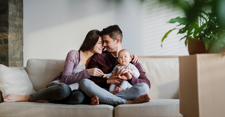 Young parents sat cuddling on their cream sofa in their livingroom whilst holding their newborn baby