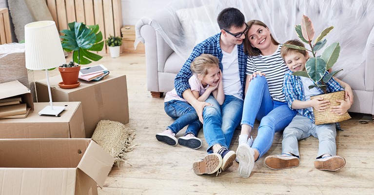 Young family of four sat their living room floor surrounded by brown boxes filled with their belongings