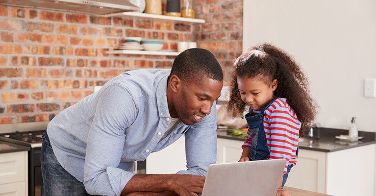 Young girl sat on the kitchen island whilst dad is with her pointing something out on his silver laptop