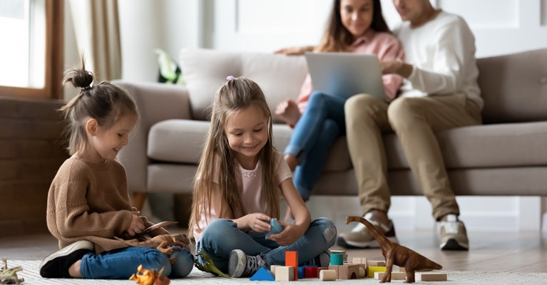 Two young children sat on the living room floor playing with their toys whilst their parents are on their laptop sat on the sofa