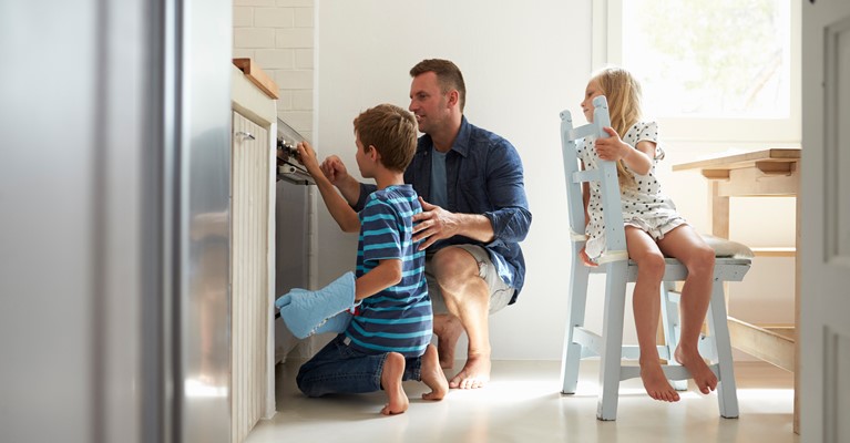 Young boy holding a blue oven glove and moving the dial on the black oven whilst dad is supervising and sister is watching