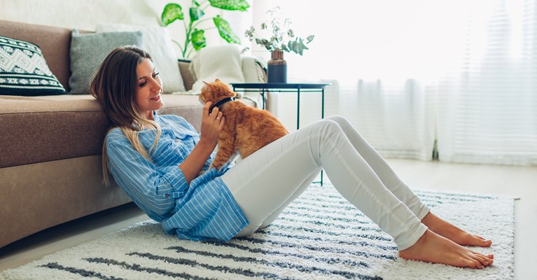 Young lady leaning against her brown sofa, stroking her ginger cat that is sat on her white jeans on a summers day