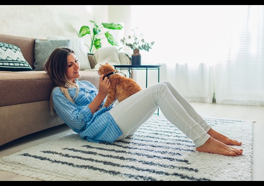 Young lady leaning against her brown sofa, stroking her ginger cat that is sat on her white jeans on a summers day