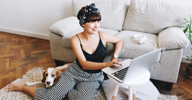 Woman sat on the floor in front of her sofa working from her laptop whilst her dog is cuddled up behind her