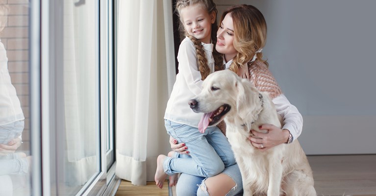 Woman holding a young girl in one arm and stroking her gold retriever in the other whilst looking out of the window