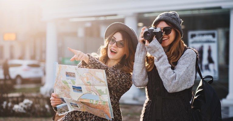 Two young girls on a city break. One pointing at something in the distance and holding a map whilst the other is taking photos