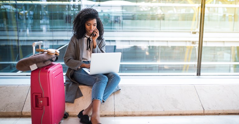 Young girl sat on the floor with her pink suitcase in an airport on her phone and laptop 