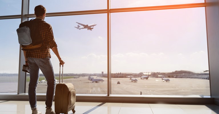 A young man in the airport with his suitcase, backpack and mobile phone in hand looking out onto the runway 