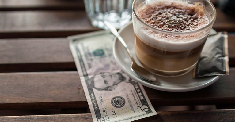 A latte in a clear coffee mug sat on a white saucer with a five dollar bill underneath 