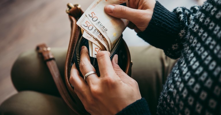 A brown crossbody bag is placed on a person's lap. A few hundred euros are being taken out of a black purse which was taken from the bag.