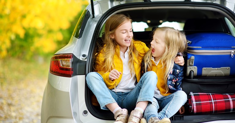 Two young girls sat with their camping luggage in the back of their car's open boot which is pulled up in a wooded area in the day