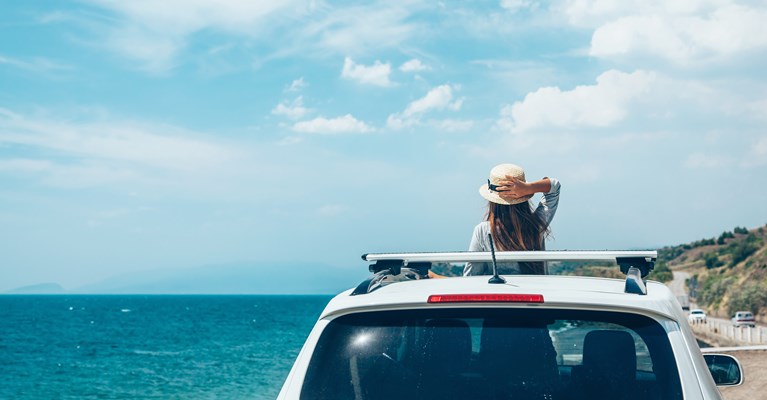 Young women holding onto her summer hat whilst stood up inside her car with her head popping out of the sunroof looking out onto the sea