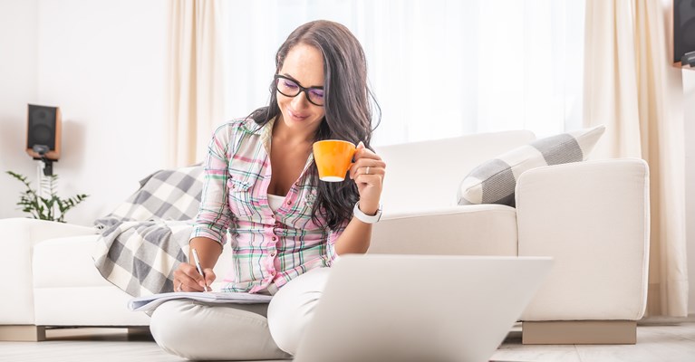 Lady sat on floor with drink and laptop taking notes in a notepad