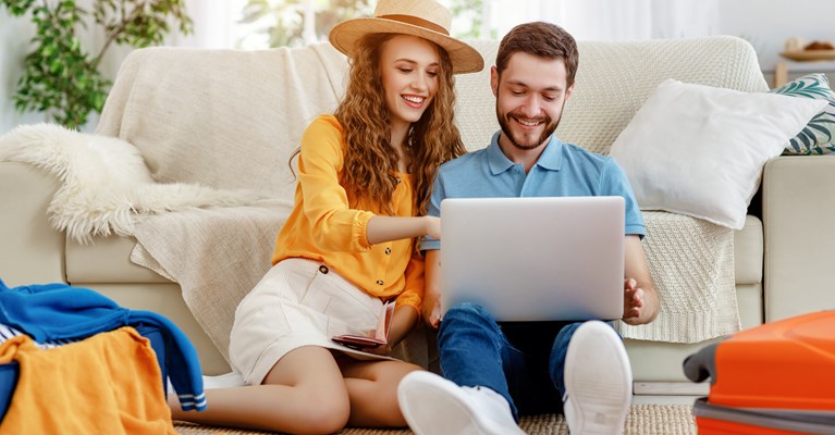 Couple sat at home with suitcase looking on laptop