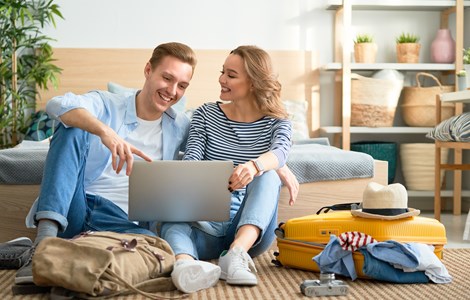 Couple sat at home with suitcase and backpack looking on laptop