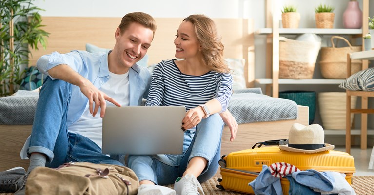 Couple sat at home with suitcase and backpack looking on laptop