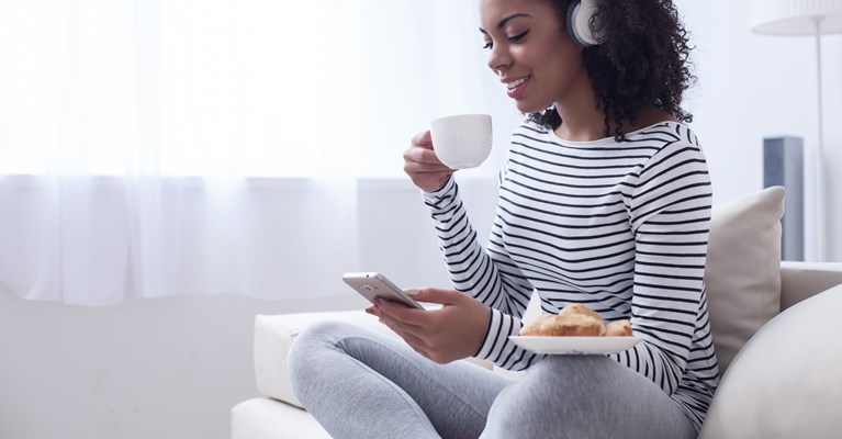 Young woman sat with hot drink and snack on her phone on the sofa