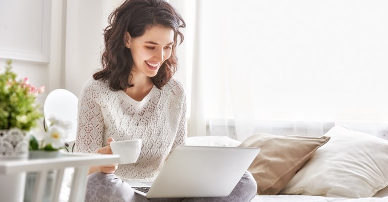 Young woman smiling with drink and laptop sat on bed