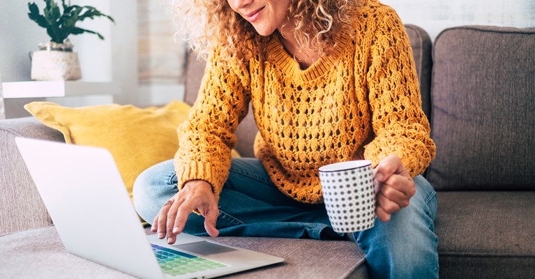 Woman sat on sofa wit a hot drink looking at laptop