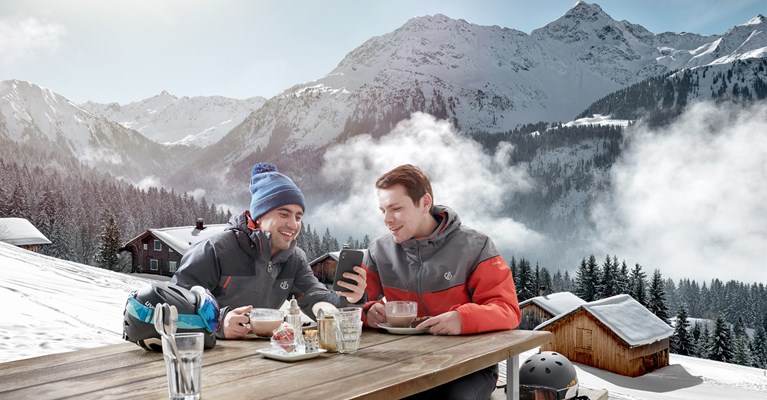 Male couple sat drinking hot drinks on mountain looking at phone