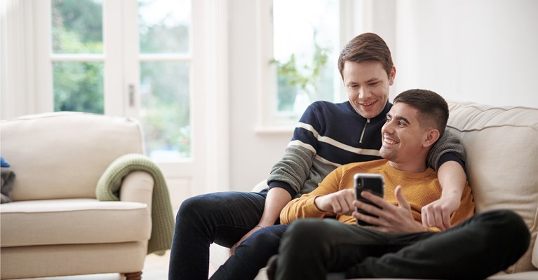Young male couple cuddled up on sofa looking at phone together