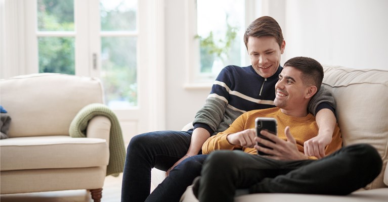 Young male couple cuddled up on sofa looking at phone together