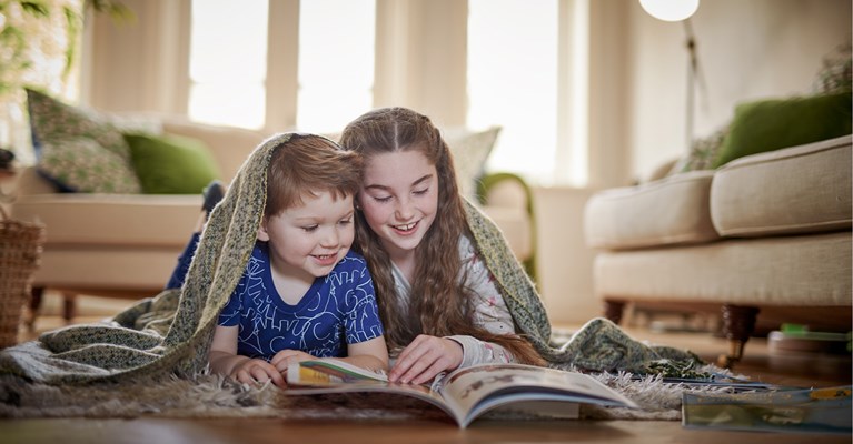 Two young children laid on the floor under a blanket reading a book together