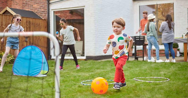 Young boy going to kick ball in the goal with family in the garden behind him