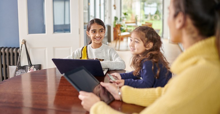 Mum and daughters sat around table looking at various devices