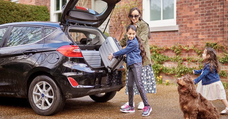 Mum loading suitcases into boot of car with daughters and dog