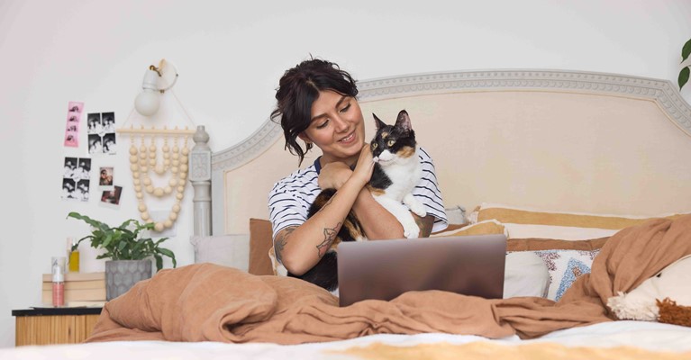 A woman looking at her laptop on her bad holding a cat
